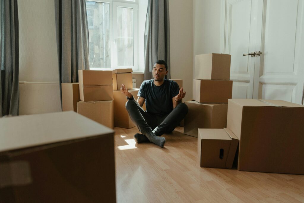 A man practices meditation surrounded by boxes in a new apartment, embracing calm during relocation.